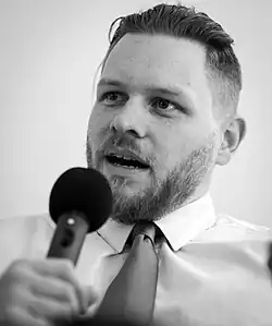 A black and white photo of Jock Palfreeman, he has light skin, a beard and is wearing a white shirt with a tie.