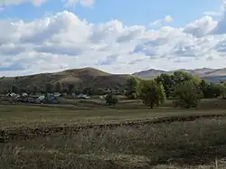 Panoramic photo depicting houses on a grassy plain with hills and a cloudy sky in the background