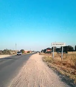 Photograph looking down a road. There is a white road sign on the right with ‘Osmanyurt’ written on it in Russian in a black font.