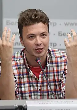 Roman Protasevich wearing a red t-shirt and white checkered shirt, seated, speaking into a tabletop microphone with both arms propped on elbows and pointed upward, gesticulating, looking right of camera