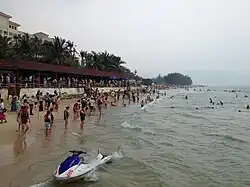 Photograph of a beach with lots of people on the sand and several in the water. There’s a jet ski in the water in the foreground.