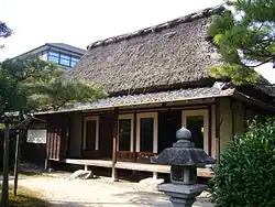 A traditional Japanese home, made entirely of wood with a thatched straw roof. A stone lantern sits in the foreground while a more modern building with glass windows can be seen in the corner of the background.