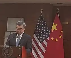 Huang Ping speaks at a lectern in front of Chinese and US flags