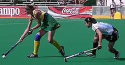 A female Australian player with blonde hair moves crouched over on the left of the screen while holding her hockey stick down low to move the ball. Behind her is a brown haired Argentinian player who is also crouched down and moving towards the Australian.
