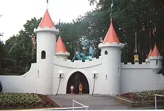 The façade of a white castle with red conical roofs, a green dragon above the gate, and two children standing in the entrance.