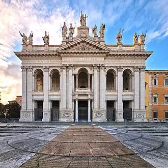 Ornate facade of the basilica with columns, main door, and statues of the twelve Apostles on the roofline, with a Latin inscription below them