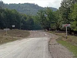 A colour photograph pointing down a road. A sign with Russian text is facing towards the camera on the right side of the road. Just past the sign, the paving of the road stops, and transitions to gravel.