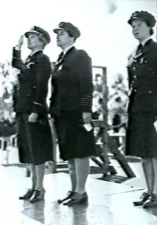 Three women in dark military uniforms standing to attention on a platform