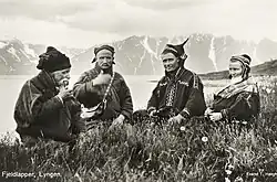 A black-and-white photograph of a group of Sámi people seated outdoors in a grassy landscape with mountains and water in the background, taken in Lyngen, Troms, Norway, in 1928.