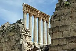 Six tall, ancient stone columns with capitals in the Corinthian style, supporting a large entablature, stand among partially ruined stone walls under a blue sky.