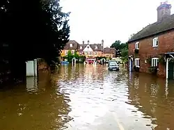 Photograph showing severe flooding to a road and housing; cars are partly submerged and fire appliances (including a dinghy) are in the distance