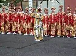 Girls in red and gold uniforms, headdresses, and white shoes stand at attention on the street behind a girl in a pastel uniform and headdress