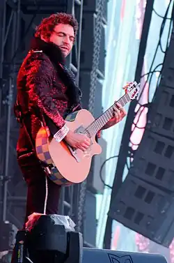 Matthieu Chedid seen from right profile, wearing a sparkling jacket and dark pants with light vertical stipe, playing an acoustic guitar on an outdoor stage, with eyes closed