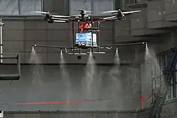 A large, multi-rotor industrial drone hovers mid-air in front of a grey building. A translucent blue tank is mounted to its undercarriage, feeding liquid into a horizontal boom that releases a wide, fine mist through multiple downward-facing nozzles.