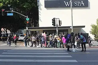Demonstrators, specifically pro-LGBTQ demonstrators, line the curb outside the black-yellow police tape line created by the LAPD, whose officers are barricading the entrance to Wi Spa. In the foreground, two demonstrators can be seen sorting beverages.