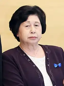 A portrait photo of a dark-haired Japanese woman wearing a light shirt under a dark sweater; she is looking into the camera with a formal expression