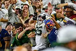 Tucker Kraft in the stands performing the Lambeau Leap with fans surrounding him.