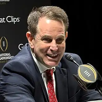 A football coach laughs in front of a microphone.
