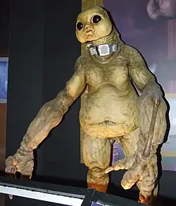 A large, green humanoid creature with bulbous black eyes. It has large, clawed hands and wears a mechanical belt-like device around its neck.
