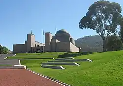 A view looking up a landscaped hill; steps lead to the entrance of a large cupola-topped building with three flag poles in front.