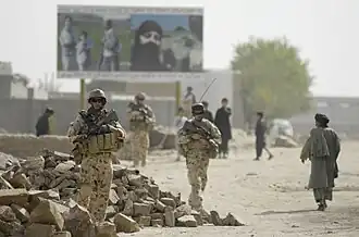 Australian soldiers from 3 RAR patrol through a sandy, rubble-strewn street in Tarin Kowt, with local civilians visible in the background, August 2008.