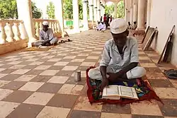A dark-skinned boy sits cross-legged on a small mat on a tiled floor in a shaded colonnade. He is wearing light clothes and a white cap and is looking down at a book written in Arabic. Beside him is a tin can. Some distance behind him are more boys sitting and standing.