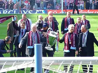 In the foreground is two men holding a large cup, they have claret scarves and a medal around their necks. Around them are ten old players in suits with medals and scarves around their necks