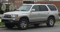 A light gray sport-utility vehicle parked on a street with brick buildings in the background, facing left