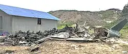 A burnt-out aircraft fuselage lying on dry ground next to a building