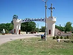 Photograph of a white structure built over a road so cars can pass underneath it. It has ‘Fernández’ written in black letters across it