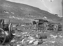 Cart wheels, wagons destroyed barrels and other wreckage in foreground, two soldiers on the road in the middle distance near a staff car