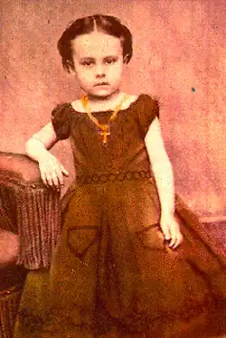 Sepia-toned studio portrait of a young girl identified as E. Pauline Johnson, standing beside a chair and wearing a dark dress with puffed sleeves and a cross necklace, her hair parted and pulled back as she faces the camera with a serious expression.