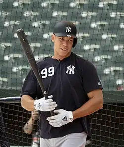 Medium-wide shot of Aaron Judge holding a bat and wearing a "NY" shirt and batting helmet.