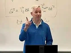 A tall man with white hair and a blue overshirt pointing up, with physics equations on a whiteboard behind him
