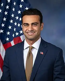 Official House portrait of Hamadeh smiling in front of the U.S. flag, wearing a black suit with American flag lapel pin, white shirt, and brown tie.