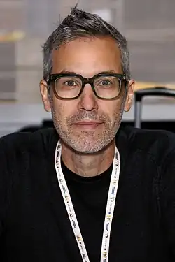 Headshot of Adam Rex with short black and gray hair and beard stubble wearing glasses and a black shirt looking at camera
