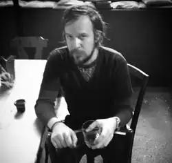 A monochromatic picture of Adam Wiltzie sitting on a chair and holding a glass cup
