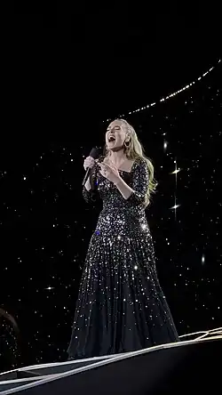 Adele sings into her microphone, dressed in a black gown ornamented with mirror shards