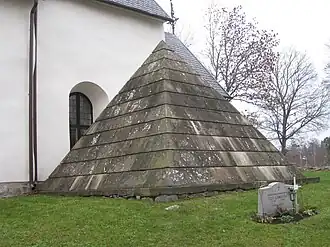 Weathered stone pyramid-shaped tomb with layered slabs leaning against white church wall, with gravestone and cross in grass nearby