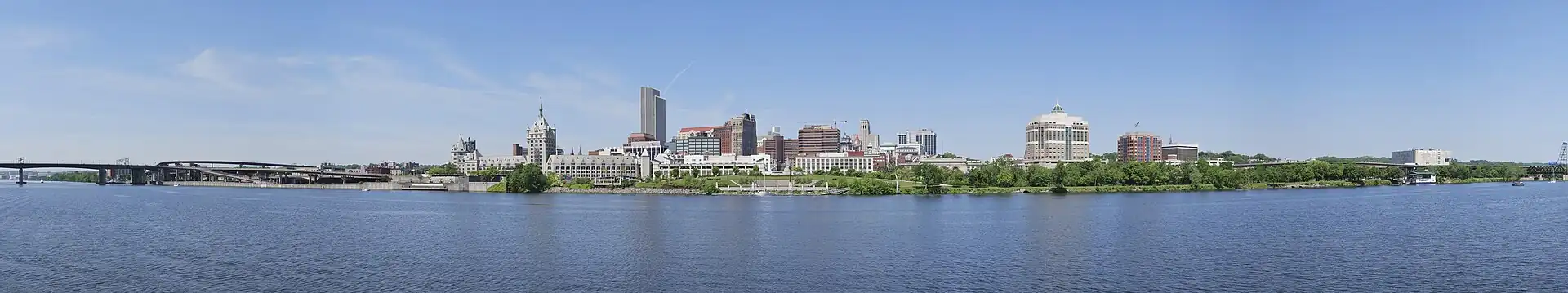 A panorama shows a river in the bottom half, crossed by a highway bridge on left; building towers are seen around the center, where a green zone on the bank of the river is seen, which extends to the right extreme of the image.