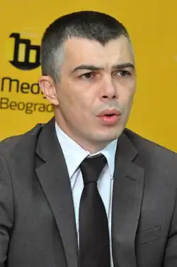 Picture of Aleksandar Jablanovic, speaking to press, wearing a suit in front of a yellow background.