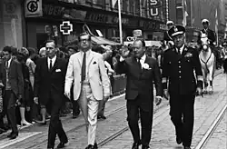 Photograph of the men in suits walking down a street with onlookers on each side. Panu Toivonen wears white. There are horse mounted patrolmen behind them on white horses.