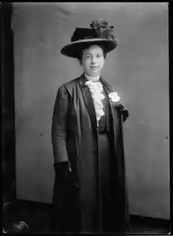 20th-century B&W portrait photo, 3/4 length, of a standing white woman who is looking directly at the came; her hair in an up-do; she is wearing a dark suit jacket, dark vest, white frilly blouse, white flower lapel, dark gloves, large dark hat.