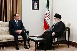 Two men sitting in discussion, one in suit, the other with white beard and turban