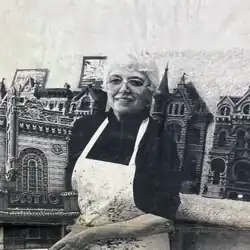 Woman wearing glasses and a paint-stained apron standing in front of detailed miniature architectural models of ornate buildings