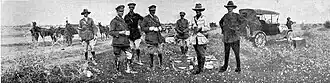 Group of officers having a cup of tea with horses and cars in the background