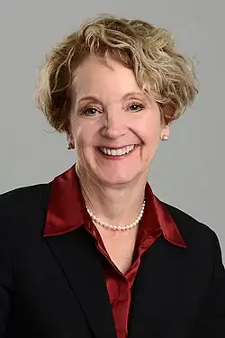 Person wearing a black blazer over a deep red satin blouse, accessorized with a pearl necklace and pearl stud earrings, posed against a plain gray background.