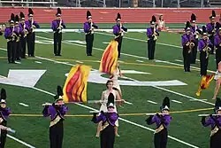 Two color guard members dressed in togas spin flags with fire designs in the middle of the DVC football field. Surrounding these color guard members are flute and saxophone players standing still and playing. They are dressed in a purple jacket and black marching pants and are wearing shakos.