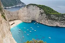 High-angle view of a turquoise bay in Zakynthos with white sand, a shipwreck, and numerous sailboats anchored between steep limestone cliffs.