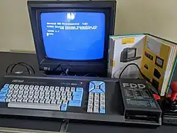 Photograph of a black home computer. The computer is a small flat rectangular single unit with a keyboard and floppy disk drive integrated onto the top. Black cables run from the back of the unit to a colour monitor behind it. The monitor reads: "Amstrad 64K microcomputer (v2). ©1984 Amstrad Consumer Electronics plc and Locomotive Software Ltd. Basic 1.1 Ready."
